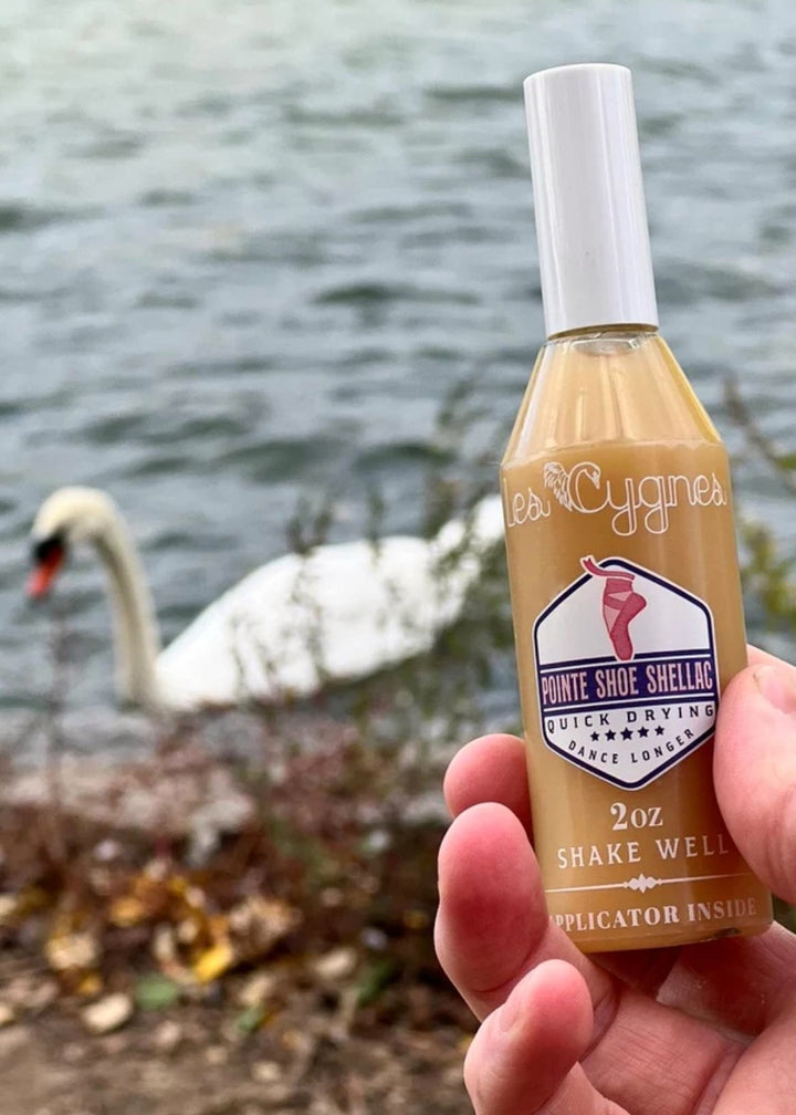 2 ounce bottle of Les Cygnes Pointe Shoe Shellac pictured next to a lake with an elegant swan floating on the water