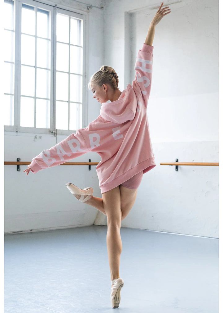 Barre Trash OH Oversized Hoodie in Ballerina Pink with white lettering, modeled by a ballet dancer en pointe in a dance studio