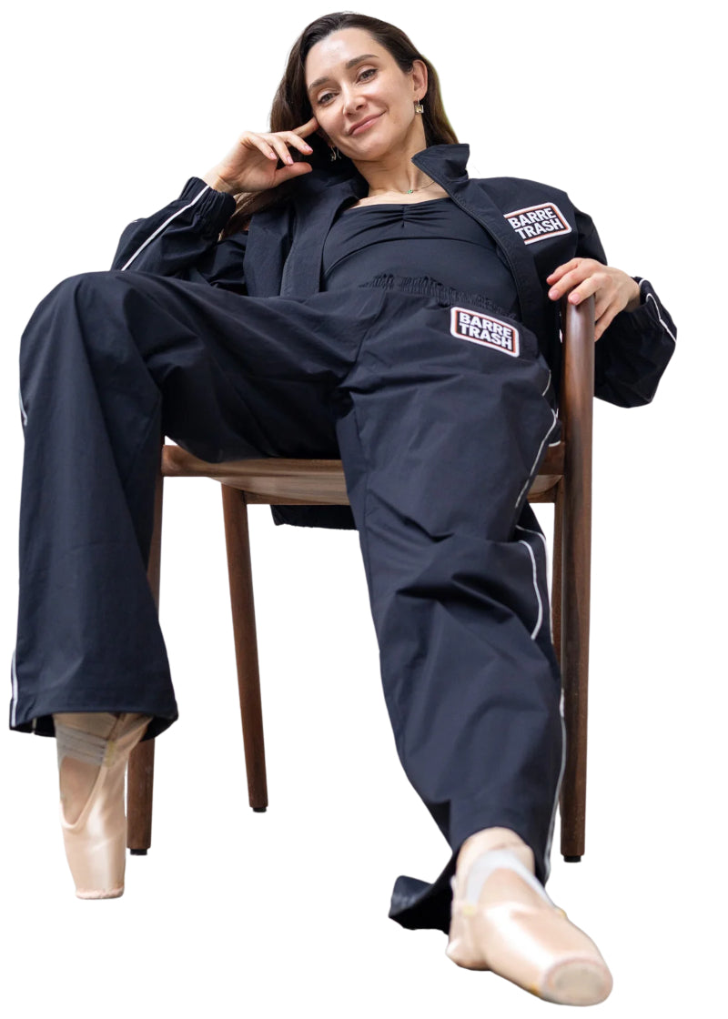 Barre Trash TSP Tracksuit Pants in Black with white side piping and "BARRE TRASH" on the left hip, modeled by a ballet dancer lounging in a chair