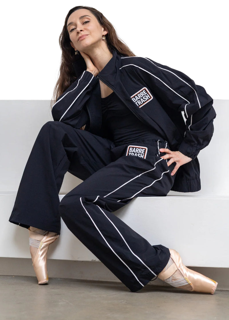 Barre Trash TSP Tracksuit Pants in Black with white side piping and "BARRE TRASH" on the left hip, modeled by a ballet dancer