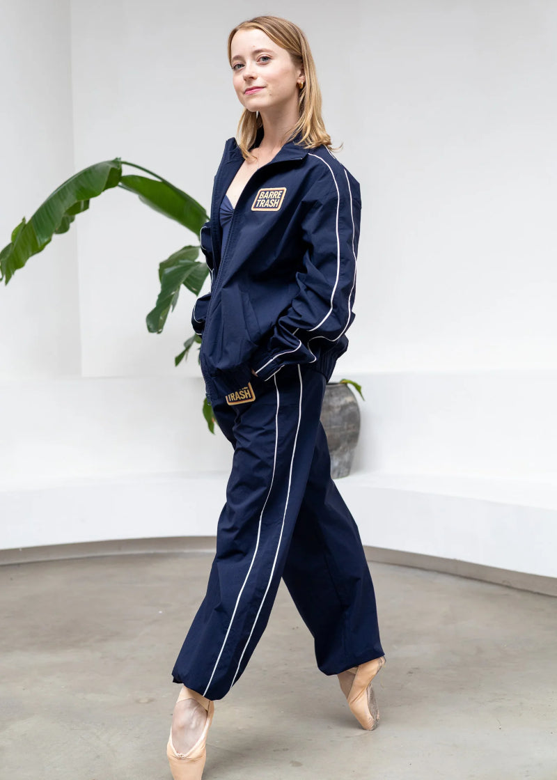 Barre Trash TSP Tracksuit Pants in Navy Blue with white side stripes and adjustable drawstring leg openings, modeled by a ballet dancer in a studio with a monstera in the background