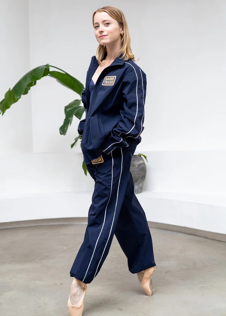 Barre Trash TSP Tracksuit Pants in Navy Blue with white side stripes and adjustable drawstring leg openings, modeled by a ballet dancer in a studio with a monstera in the background